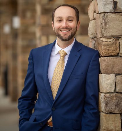 Person wearing a blue suit jacket, white shirt, and patterned yellow tie standing outdoors beside a stone wall. The person is posed slightly angled with hands in pockets, and the background shows additional stone columns out of focus.